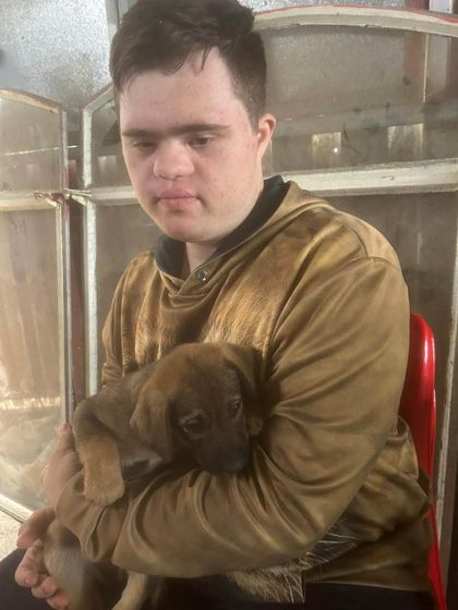 Young man in a tan jacket holding a brown puppy while seated indoors