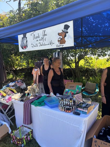 Two women stand behind a table of donated goods under a blue tent with a charity banner overhead