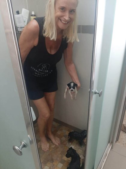 A smiling volunteer in an SSWC top holds a puppy in a shower, with more puppies at her feet