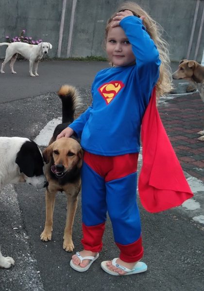 A little girl in a Superman costume with a red cape pats a brown stray dog on a street, with other dogs around her