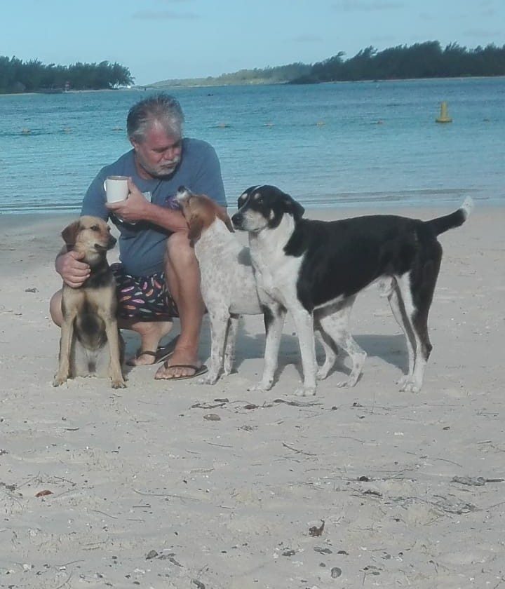 Princess, a small brown dog, sits beside a man holding a mug on Blue Bay beach, next to two other beach dogs, Jingo and Pepe