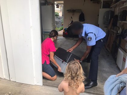 A police officer in uniform and the vet lift a dog carrier into the garage while a toddler watches from behind