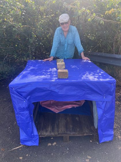 A smiling man in a cap leans on the finished shelter, its blue tarp weighed down with three bricks